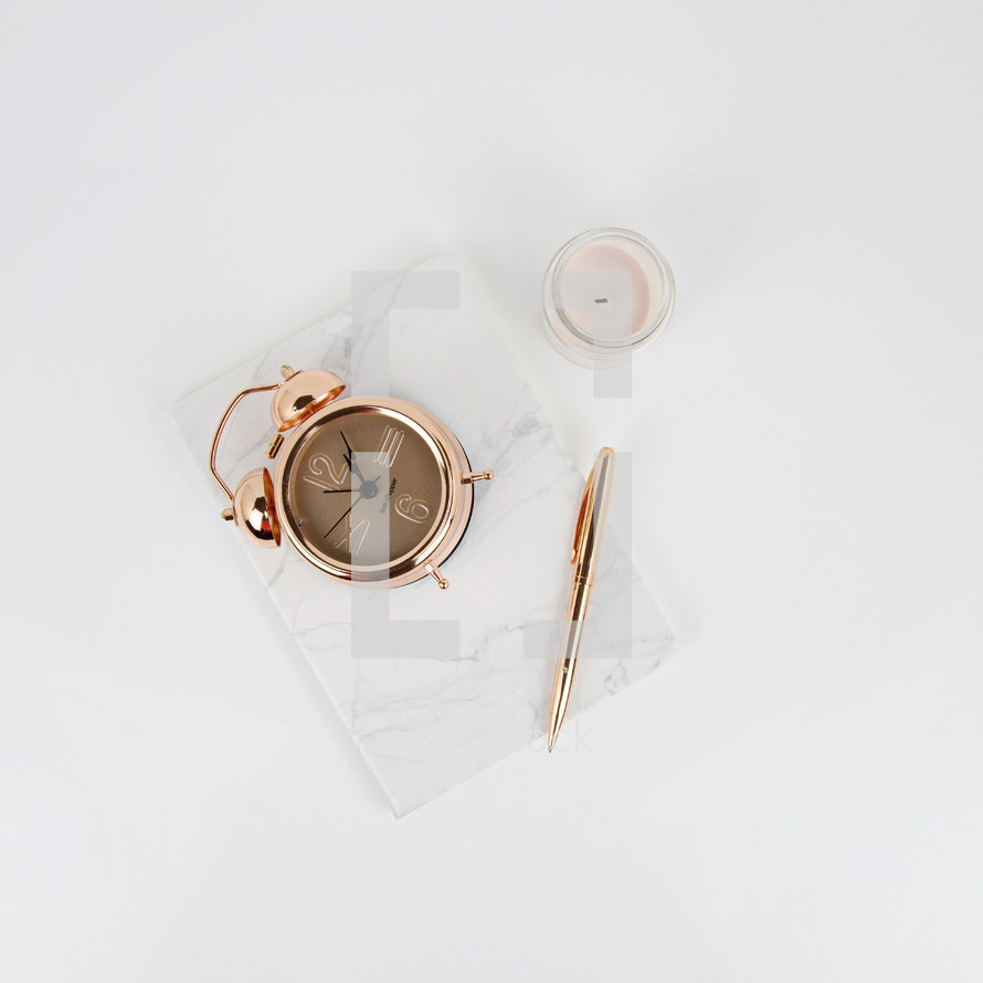 rose gold, alarm clock, flower, notebook, pen, and candle on white desk 