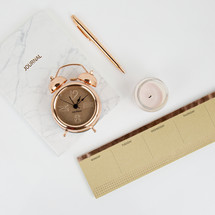 rose gold, alarm clock, calendar, candle, journal, pen on white desk