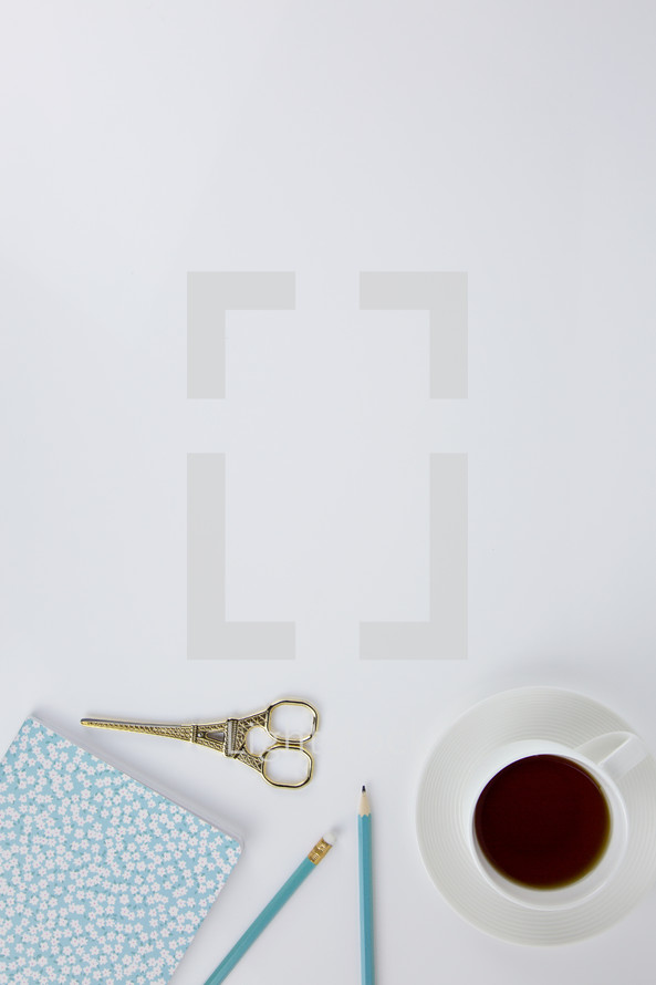 gold scissors, floral notebook, blue pencils, and coffee cup on white background 