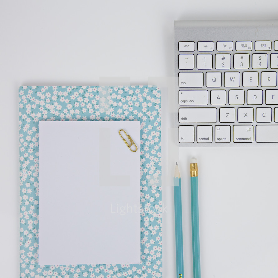 floral notebook, paperclip, blue pencils, and computer keyboard on a desk 