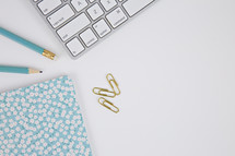 gold paperclips, computer keyboard, floral planner, and blue pencils on a white background 