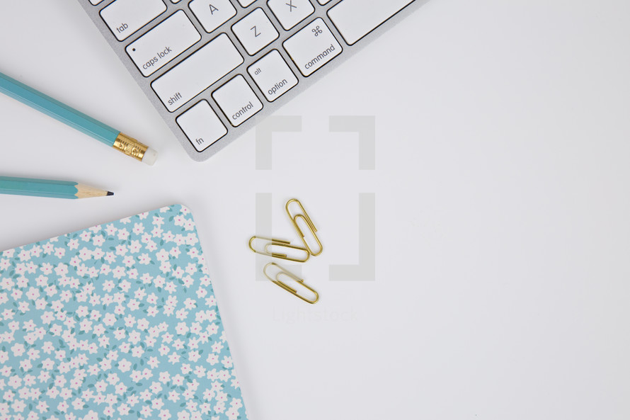 gold paperclips, computer keyboard, floral planner, and blue pencils on a white background 