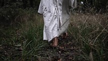 The feet of Jesus Christ, or biblical, spiritual prophet in white, tattered robe walking in dramatic slow motion in woods surrounded by trees.
