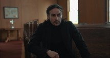 Young, emotional, anxious, and stressed man with long hair and black suit sitting in old church in worship and praying.