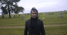 Sombre, sad young man in black suit walking in cinematic slow motion in cemetery through graveyard tombstones.