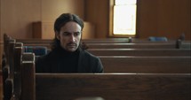 Young, emotional, anxious, and stressed man with long hair and black suit sitting in old church in worship and praying.