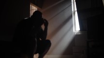 Depressed, sad and grieving man crying and praying holding his face in his hands illuminated by light coming through window.