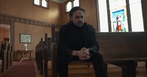 Young man praying in old church building sitting in pew with stained glass church window behind him.