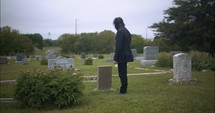 Young, sad man in black suit in cemetery at graveyard tombstone grieving in cinematic slow motion.