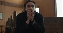 Young, emotional, anxious, and stressed man with long hair and black suit sitting in old church in worship and praying.