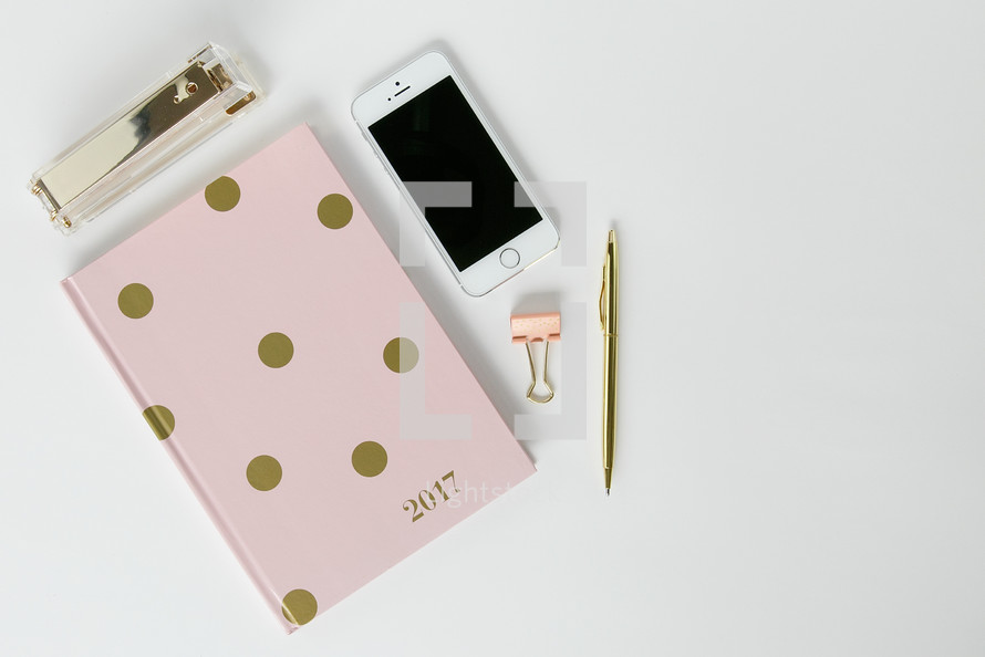 pink and gold polka dot planner, stapler, clip, pen, and cellphone on a desk 