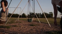 A father pushes his small, happy child in the sunset or sunrise sunlight on a swing at a park in cinematic slow motion on Father's day.