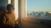 A 12-year-old boy at an urban window during sunrise, wearing white headphones and singing along to his phone