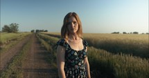 Young woman on dirt road wearing flower dress looking at camera.