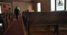 Young man in black suit walking into old church building with stained glass window sits down on pew.