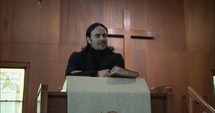 Young pastor at pulpit in black suit in old church building preaching, teaching sermon to congregation on Sunday morning worship service in cinematic slow motion in front of wood cross.