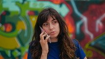 a woman talking on a cellphone standing in front of a graffiti covered wall 