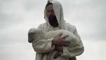 Jesus Christ, the good shepherd of Psalm 23, holding a baby lamb or sheep in his arms while wearing a white, tattered tunic and hood outside in front of cloudy sky
