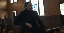 Young, emotional, anxious, and stressed man with long hair and black suit sitting in old church in worship and praying.