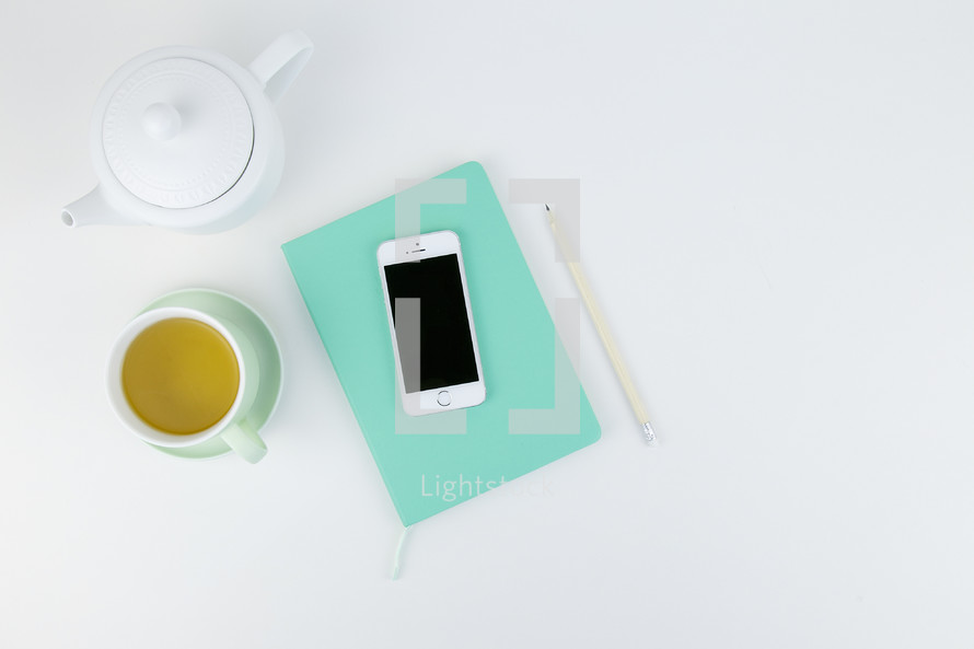 cellphone, tea pot, tea, hot tea, saucer, journal, and pencil on a white background, table