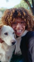Young curly-haired Latin woman embracing adorable dog outdoors, happily smiling and chatting on camera during video call. Vertical format, POV shot
