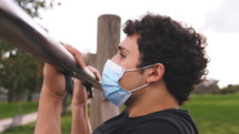 Side view of a man doing pull ups at a park wearing a medical mask.