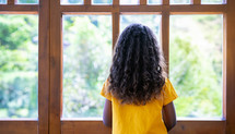 the back of a young girl looking out a window on a bright, sunny day 