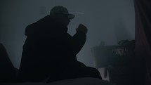 Silhouette of man wearing a hat and hooded coat in a dark, smoky room smoking a cigarette or drugs like marijuana. 