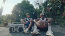 Two Basketball Friends Sitting with Ball on Playground
