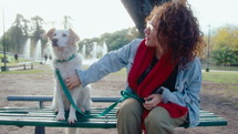 Loving female owner sitting with cute mixed-breed dog on bench in the park, petting and kissing him. Medium long shot, fountains in the background
