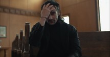 Young, emotional, anxious, and stressed man with long hair and black suit sitting in old church in worship and praying.