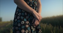 Woman wearing flower dress standing outside in grass field with crossed hands.