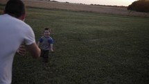A young, cute happy boy in a blue shirt running in sunrise or sunset to his father in sunlight in green grass in cinematic slow motion.