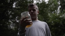 A sad, depressed young man in the woods holding a jar of alcohol.