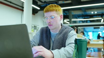 Young gen Z man wearing glasses and casual outfit sitting at desk with coffee tumbler, typing on laptop, working in open space office. Zoom out shot
