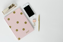 pink and gold polka dot planner, stapler, clip, pen, and cellphone on a desk 