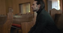 Young, emotional, anxious, and stressed man with long hair and black suit sitting in old church in worship and praying.