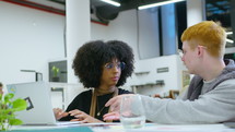 Young multi-ethnic male and female coworkers sitting at desk in open space office, working on laptop, having discussion about business project
