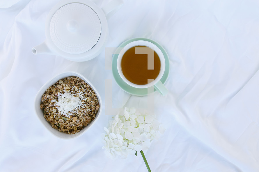 bowl of oatmeal, tea pot, cup of tea, and flowers on sheets on a bed 