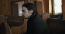 Young, emotional, anxious, and stressed man with long hair and black suit sitting in old church in worship and praying.