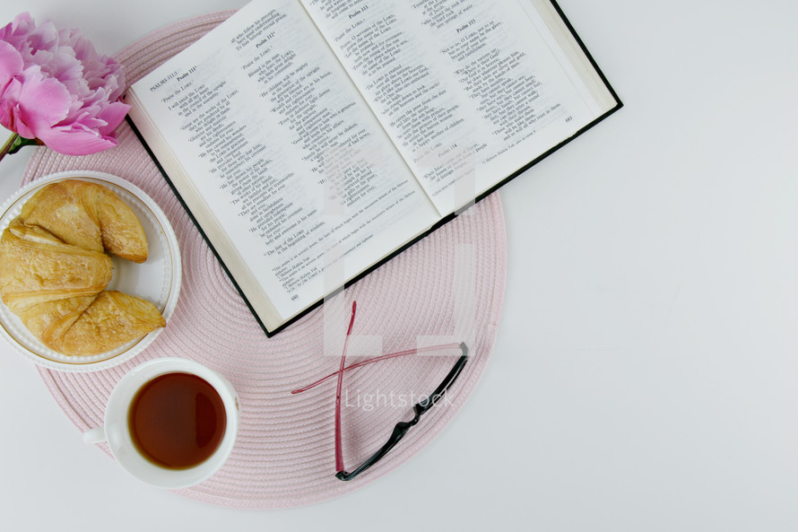 reading glasses, open Bible, tea cup, croissant, and place mat 