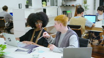 Young African-American businesswoman and Caucasian businessman sitting with laptops and papers at desk in modern open space office, discussing plans and ideas, working in team
