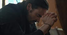 Young, emotional, anxious, and stressed man with long hair and black suit sitting in old church in worship and praying.
