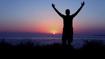Silhouette of a man raising his hands in worship and praise by the ocean at sunset.