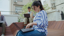 Young brunette girl in casual clothing sitting outside on bench, reading notes and typing on laptop, working or studying online on summer day in the city
