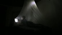a man in a tent under a mosquito net typing on a laptop 