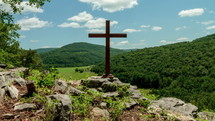 Timelapse of cloud movement over a mountain range and a wooden cross on a hill of rocks.