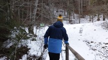 Back View Of A Person Walking On Snow Covered Wooden Bridge In Dense Forest During Winter. Dolly Shot