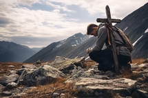 Man kneels with cross on back to pray in mountains