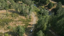 Drone shot of truck driving down a gravel road by a river on a sunny morning.